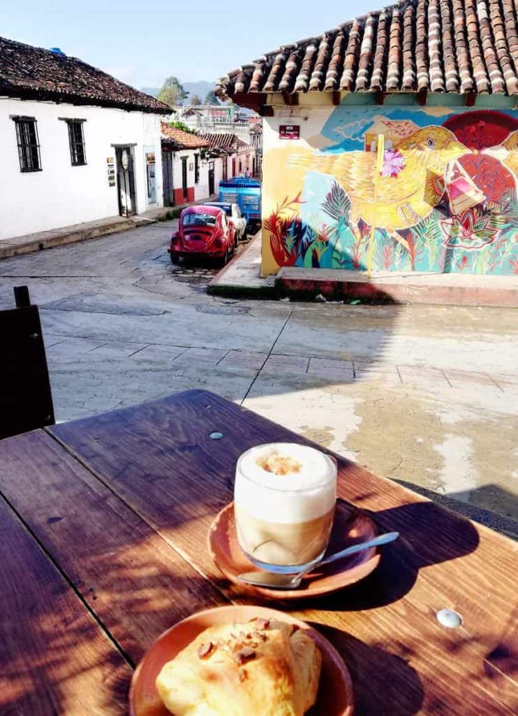 At an outdoor table overlooking a picturesque street in San Cristobal, sits a croissant and cappuccino from Kulkulpan Cafe. On a wall in the background is a colorful bird mural.