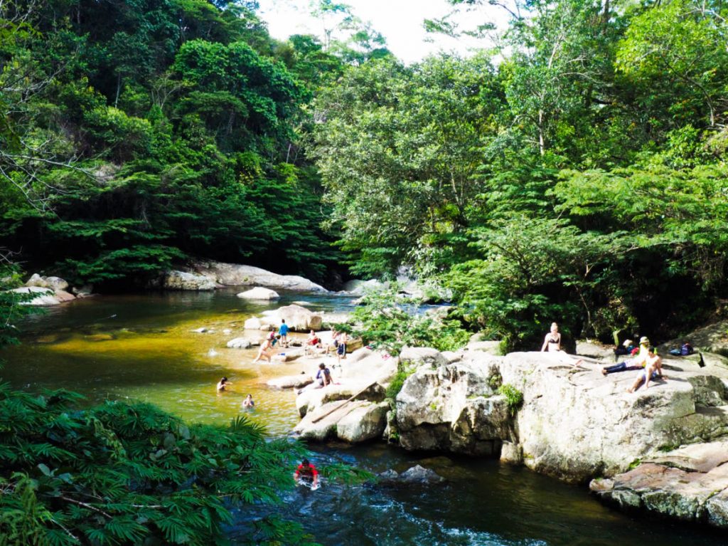 From a small bridge looking down across the river below, people play in the water and sun bathe on the large rocks surrounding the river.