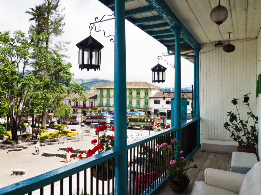 Past the bright blue columns of this second story balcony, a man walks in the square below. In the distance, other colorful balconies line the square while chiva buses wait to depart to neighboring towns.