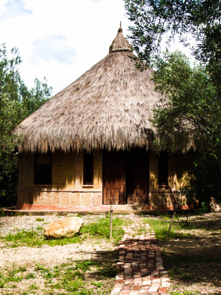 A stone pathway leads to a round clay house with thatched roof, traditional of the Muisca people.