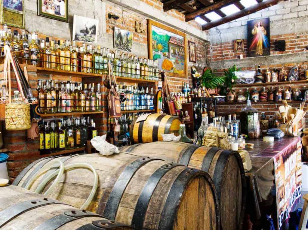 Barrels of mezcal sit to the side of the bar with shelves of mezcal bottles in the back ground at Rancho Blanco on a mezcal tour in Oaxaca.