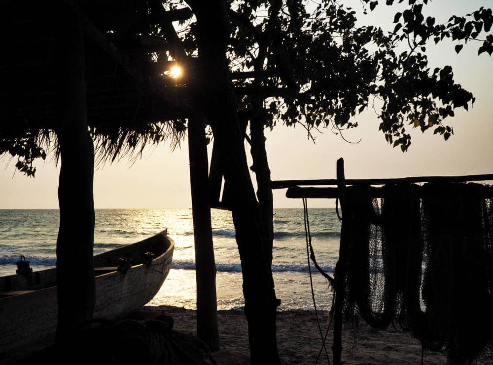 Silhouetted by the sun, a boat sits in the sand next to fishing nets hanging from a post.