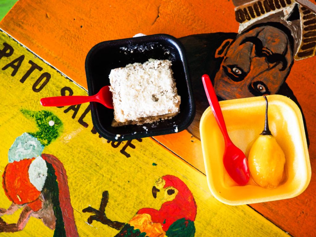 From above, on a colorful hand-painted table, two square styrofoam containers hold a dessert and a topping from San Antonio de Pereira.