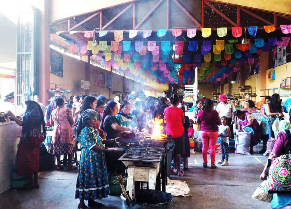 Women, many dressed in traditional clothing, gather around to cook food on the grills set up in the Tlacolula Market.